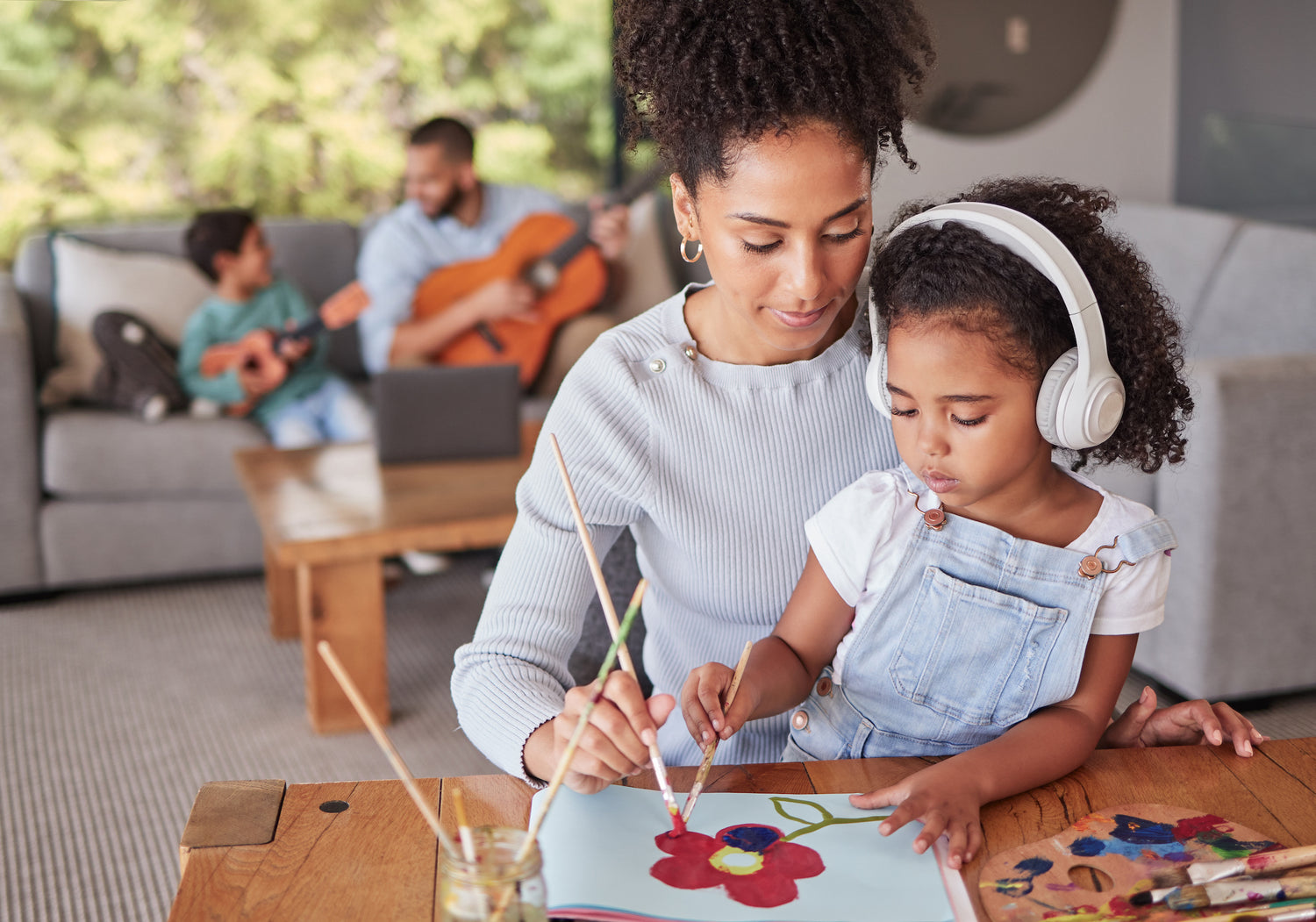 Family bonding together in the living room, dad teaching son how to play guitar and mom teaching daughter how to paint.